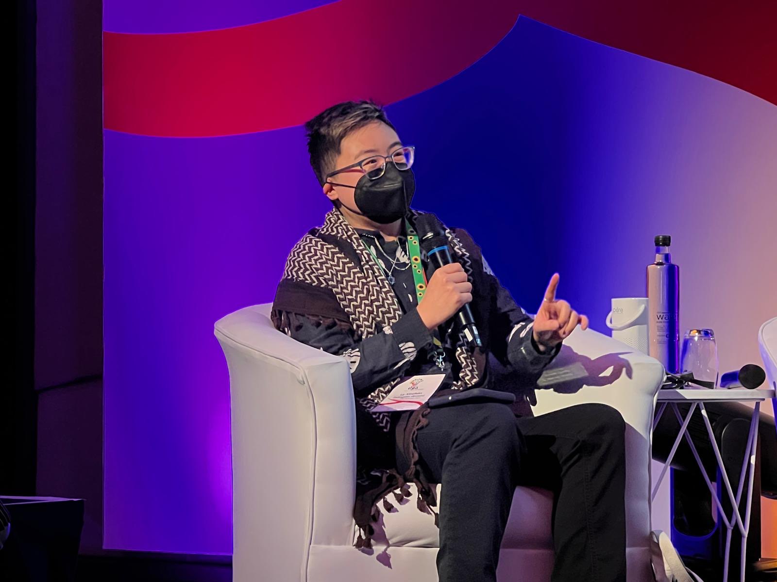 Image of Ly Xīnzhèn wearing a brown keffiyeh and a KN95 face mask, speaking into a microphone while seated on a stage.
